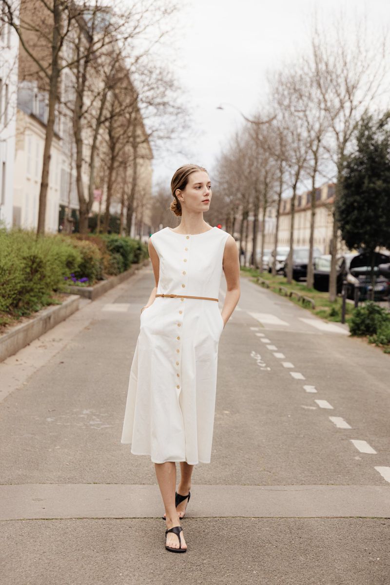 Model in a white sleeveless button-down dress walking along an urban street lined with bare trees.