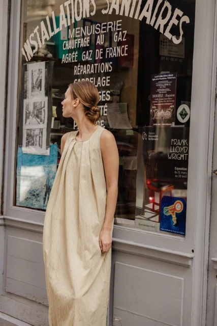 Woman in a tan sleeveless dress standing in front of a shop window with French writing. Fashion editorial in an urban setting.
