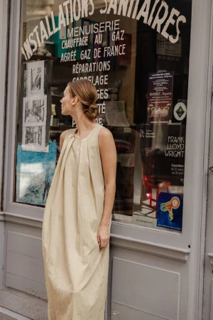 Woman in a tan sleeveless dress standing in front of a shop window with French writing. Fashion editorial in an urban setting.