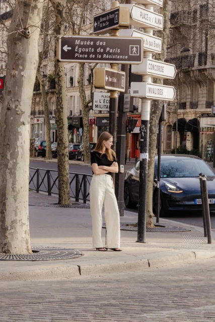 A woman stands on a cobblestone street in Paris, near street signs, wearing a black top and white pants on a sunny day.