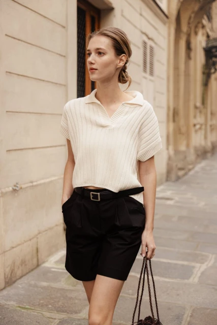 Adult woman in a polo-style top and black shorts walking outdoors against a stone building. Daytime shot, medium framing.