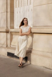 Model leaning against a pale stone building wearing a cream-colored top and skirt set and black sandals