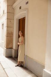 Model leaning against a wooden doorway on a white building exterior wearing a beige dress and black sandals.