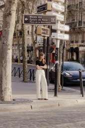A woman standing on a Parisian street corner beneath vintage street signs with cars passing on the road behind.