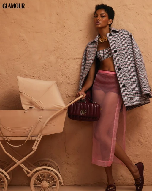 Fashion editorial shot of a woman in a plaid jacket, crop top and sheer skirt, posing with a pram and Gucci purse against a tan backdrop.