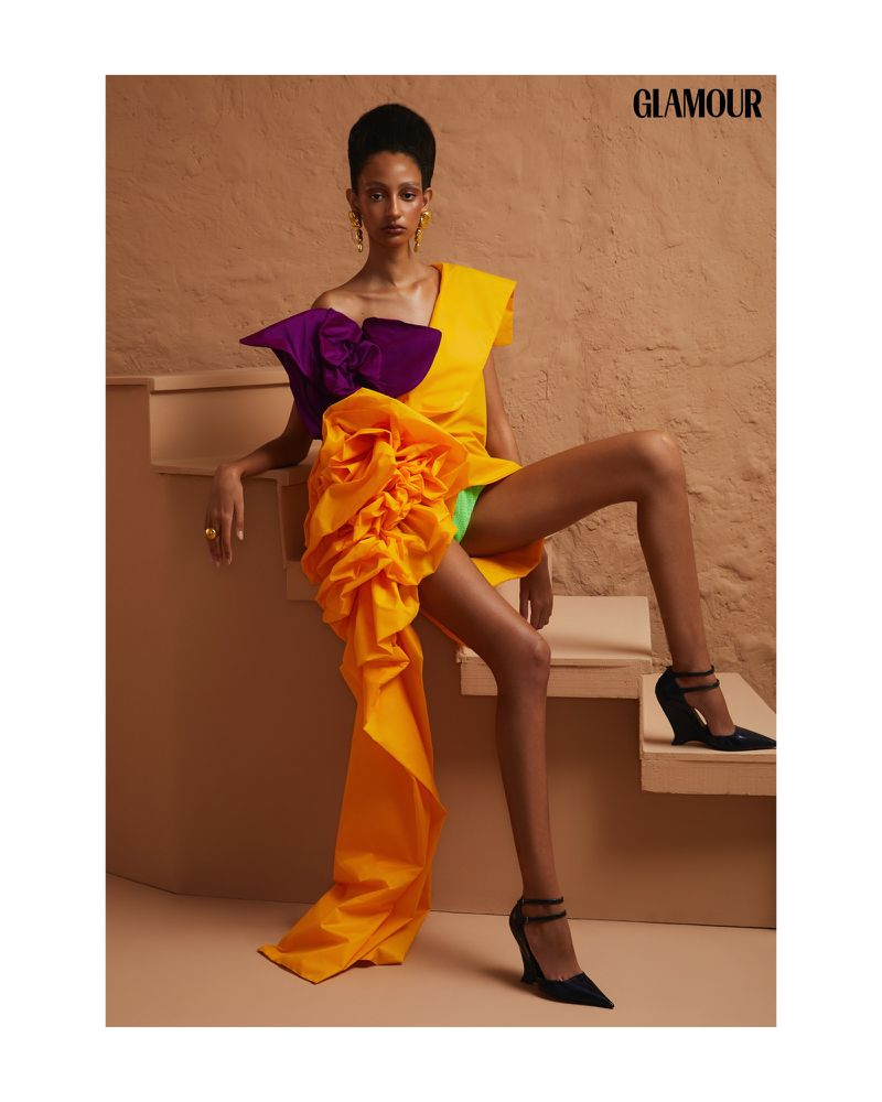 Fashion editorial shot of a young Black woman in an orange, yellow, and purple dress with black high heels posing on stairs indoors.