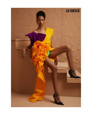 Fashion editorial shot of a young Black woman in an orange, yellow, and purple dress with black high heels posing on stairs indoors.