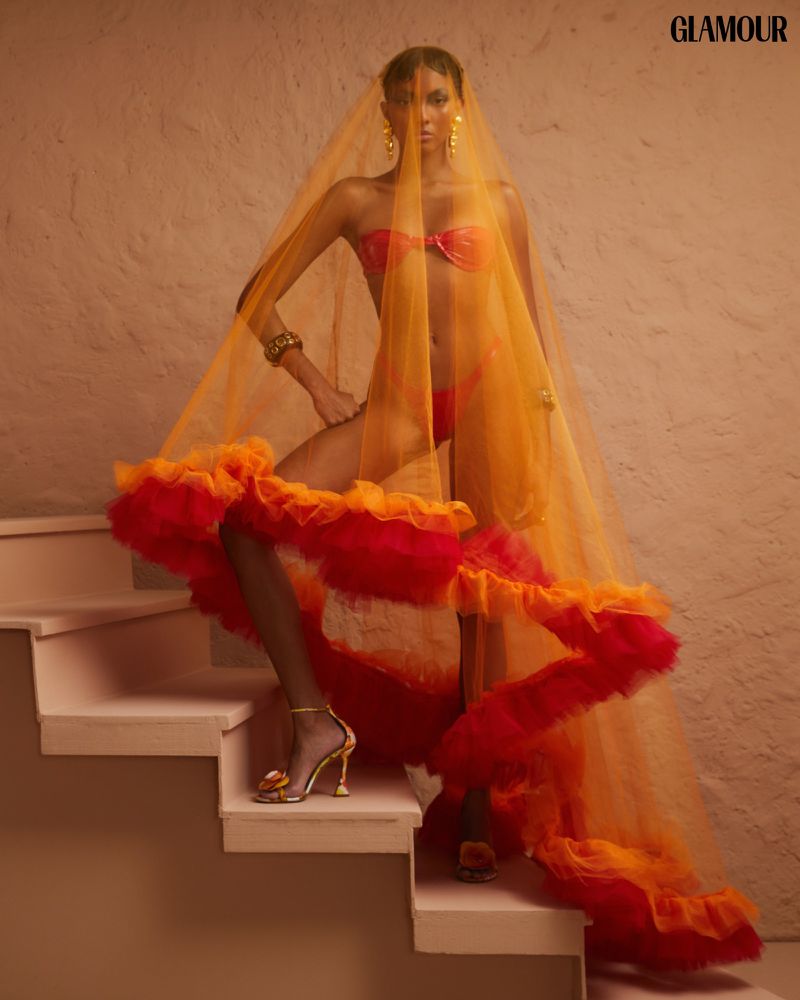 Woman in orange bikini and sheer ruffle veil poses on stairs. Fashion editorial shot with shallow depth of field.