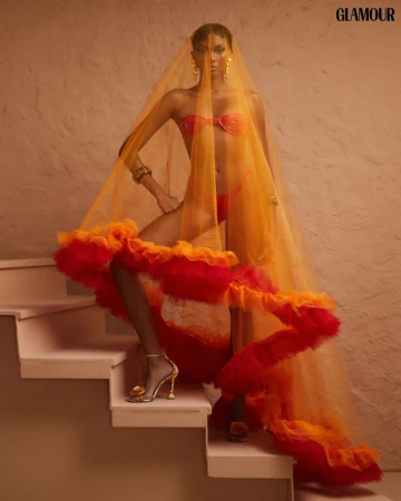 Woman in orange bikini and sheer ruffle veil poses on stairs. Fashion editorial shot with shallow depth of field.