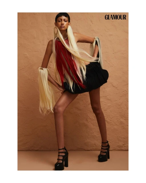 A model poses in a fashion shot wearing a black dress with red and blonde extensions and platform heels against a peach background.