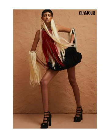A model poses in a fashion shot wearing a black dress with red and blonde extensions and platform heels against a peach background.