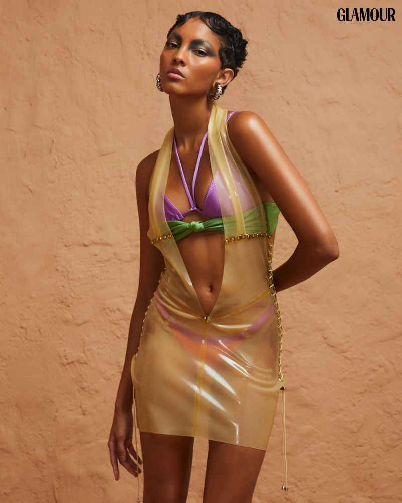 Fashion editorial shot featuring an adult female model in a yellow mini dress, posing in a studio setting against a textured wall backdrop.
