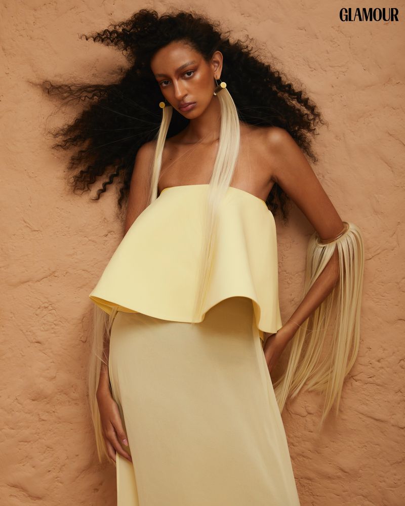 Woman in a yellow strapless dress with long fringe earrings poses against a textured backdrop, glamour editorial fashion shot.