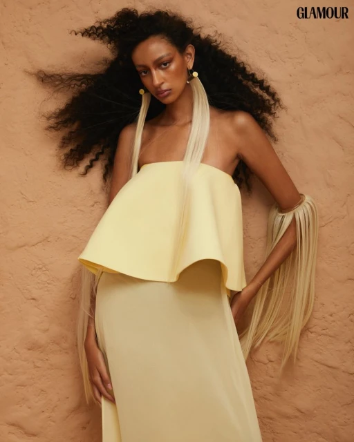 Woman in a yellow strapless dress with long fringe earrings poses against a textured backdrop, glamour editorial fashion shot.