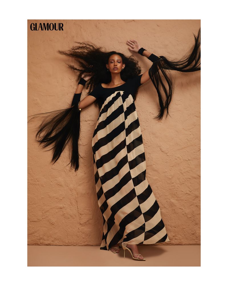 Fashion editorial shot of a Black woman in a striped dress with long flowing hair extensions against a textured beige background.