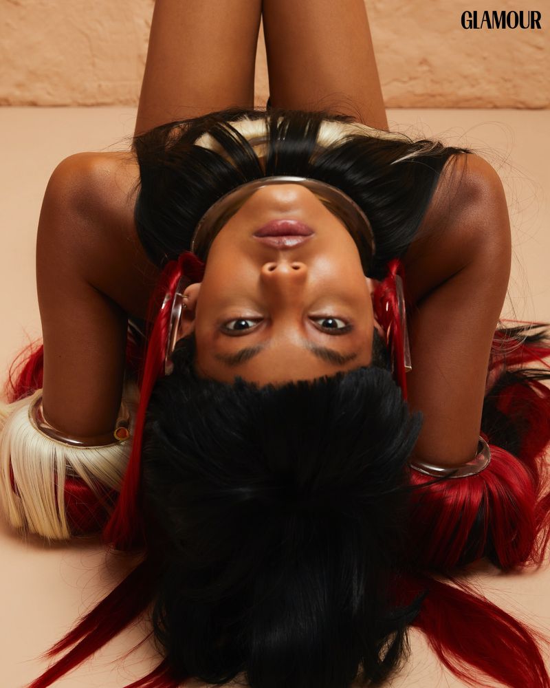 Glamour fashion editorial featuring a dark-skinned adult woman with distinctive hairstyle and jewelry against a tan background.