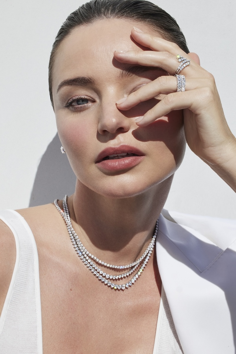 Glamorous portrait of a woman wearing a white blazer and a tiered diamond necklace with rings, posing against a white background.