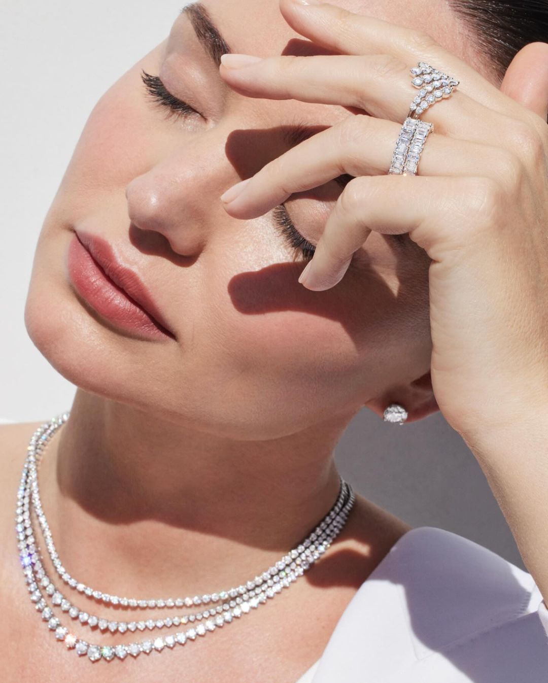 Close-up shot of a woman with her eyes closed wearing a diamond necklace, ring, and earrings in a bright, neutral indoor setting.