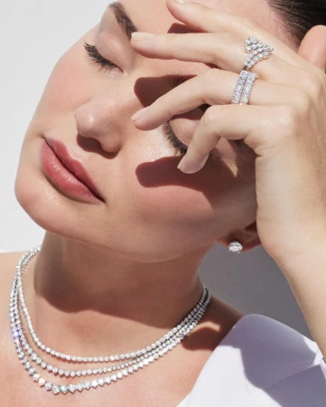 Close-up shot of a woman with her eyes closed wearing a diamond necklace, ring, and earrings in a bright, neutral indoor setting.