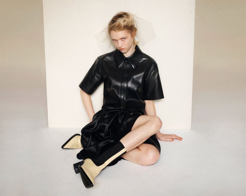 A fair-skinned model in a black leather dress and boots poses against a white backdrop, with a neutral expression and hair styled with a white net.