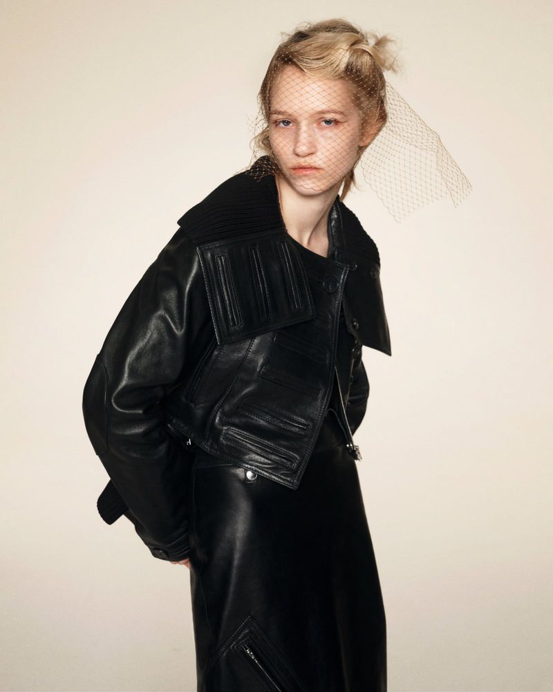 Portrait of a fair-skinned model wearing a black leather jacket, black dress, and a netted veil against an off-white background.