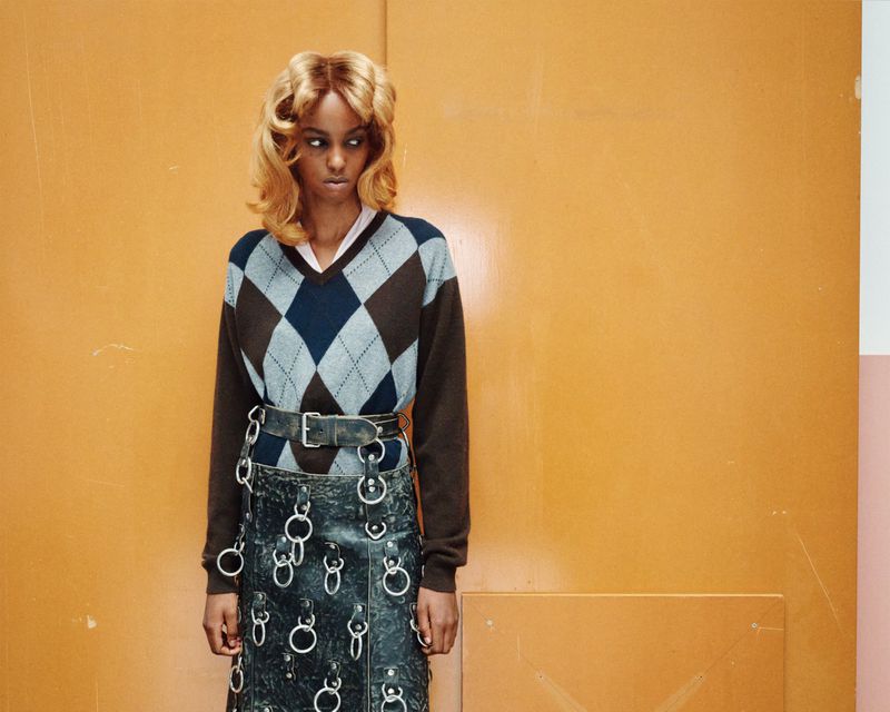 A young woman with an argyle sweater and a metal ring skirt stands against an orange and pink wall in a studio setting.