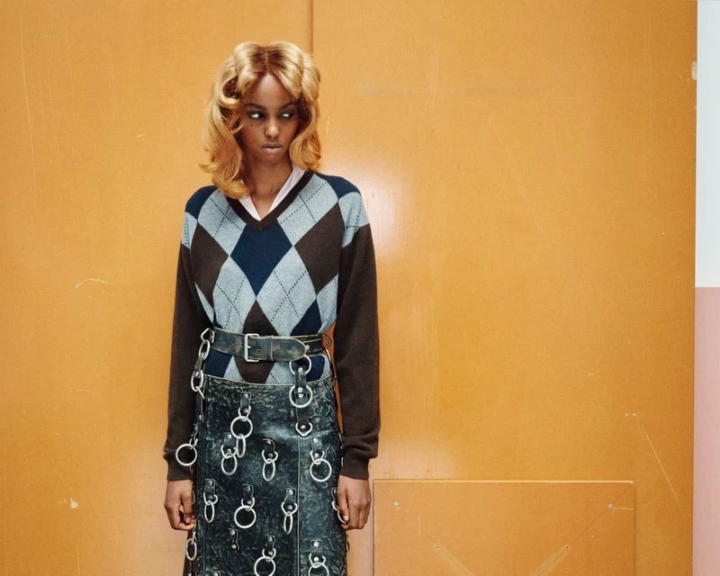 A young woman with an argyle sweater and a metal ring skirt stands against an orange and pink wall in a studio setting.