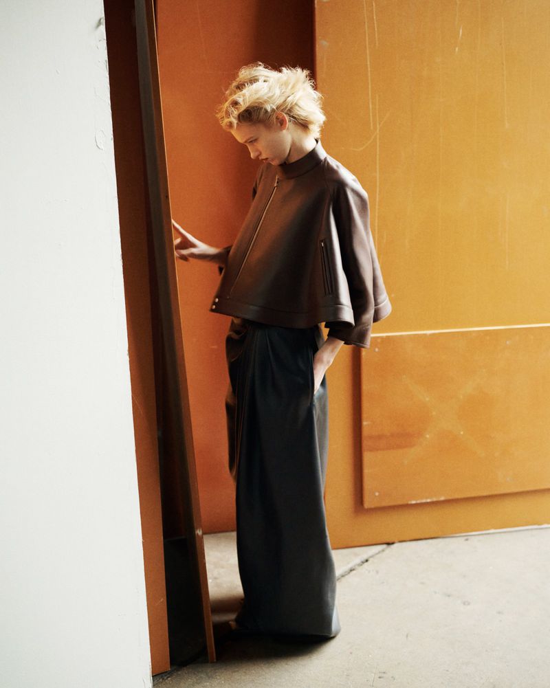 A young woman in a stylish brown leather jacket and dark, draped pants stands near a wall, side view, in a fashion editorial setting.