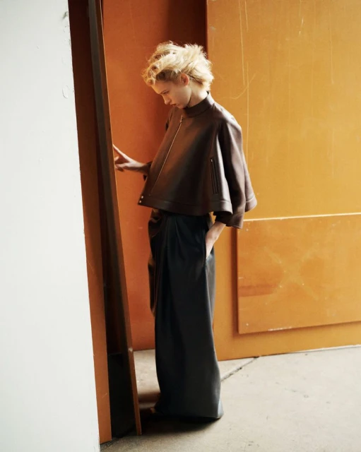 A young woman in a stylish brown leather jacket and dark, draped pants stands near a wall, side view, in a fashion editorial setting.