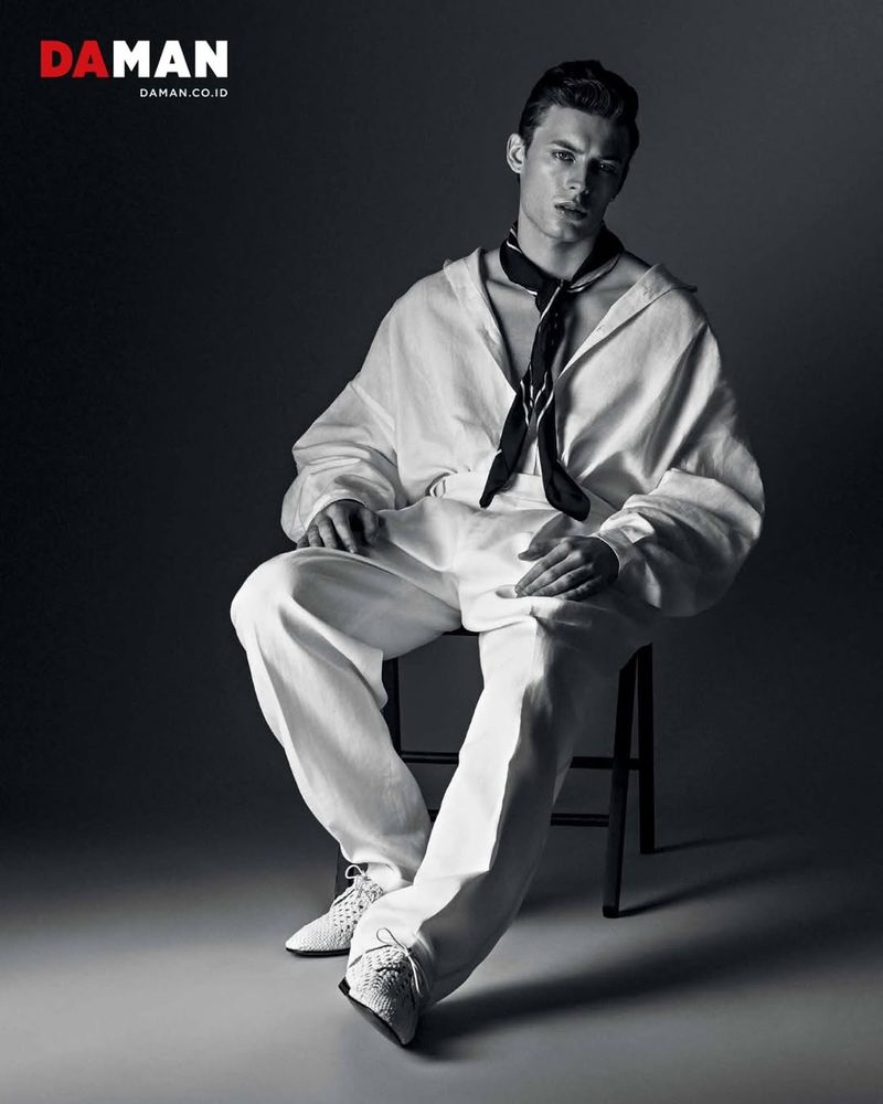Man in white sailor outfit seated against a dark background in a studio, fashion editorial shot for DA MAN magazine, stylish and elegant.