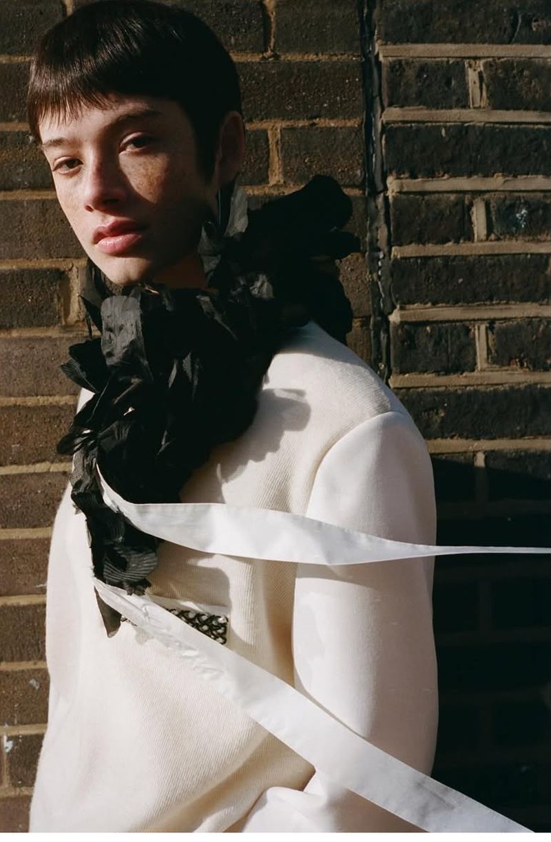 Portrait of a person with short dark hair, wearing a white coat and black decorative collar, standing against a brick wall.