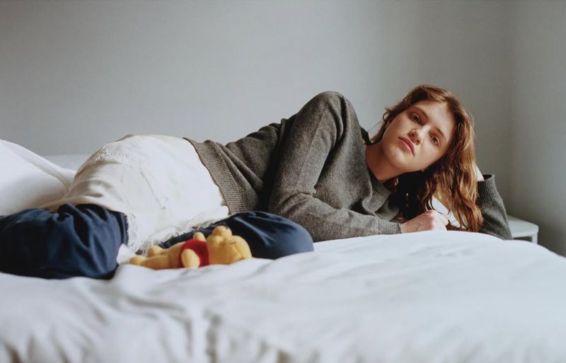 Adult female lying on a bed in a gray sweater and white skirt with a plush toy indoors, resting and looking at the camera.