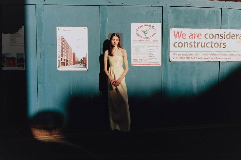Young adult woman in pale yellow dress stands against a blue wall with posters in urban setting under natural light and shadow.