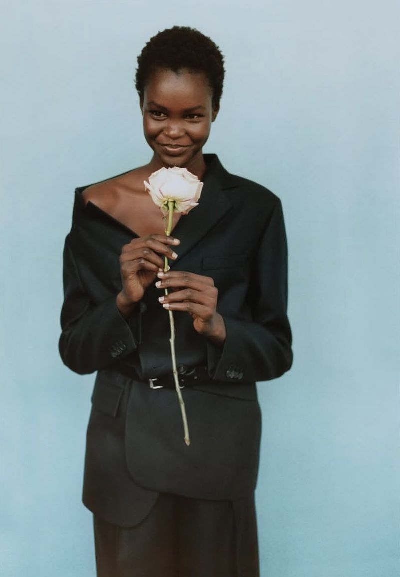 Black woman in a black blazer holding a single white rose against a plain light blue background.