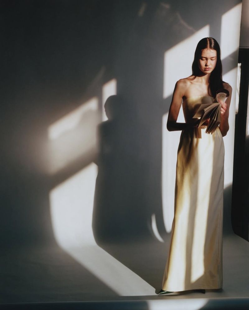 Model holding a book in a long cream dress against a grey studio backdrop with dramatic rectangular light and shadow patterns.