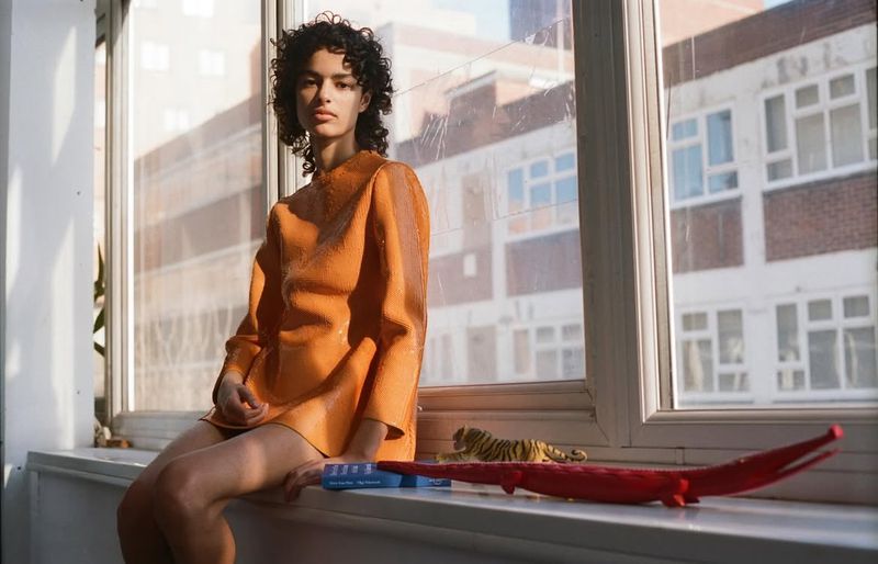 A woman in an orange dress sits on a window sill inside an apartment with toy animals alongside a window and a cityscape view.