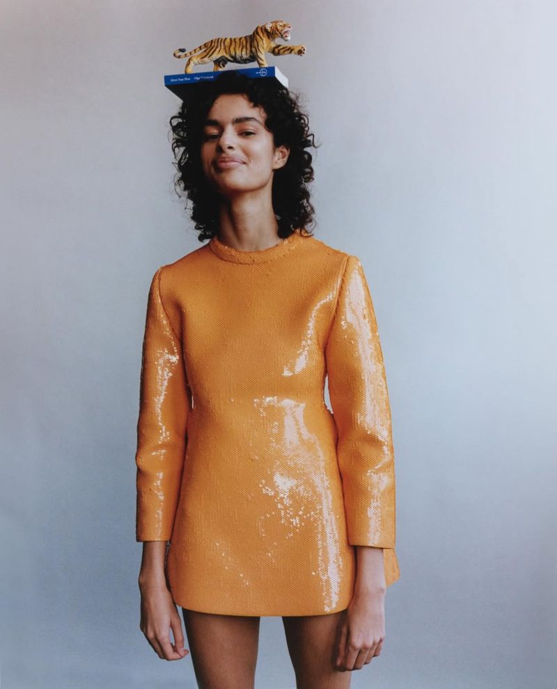A woman in an orange dress balances a tiger figurine on a book atop her head in a fashion forward and playful studio portrait.