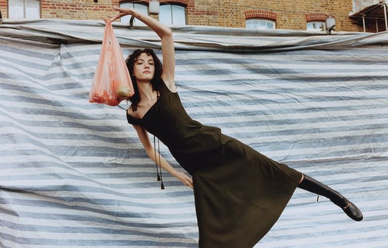 A woman in a dress holds a plastic bag of fruit against a striped backdrop and brick building in an outdoor setting, fashion style.