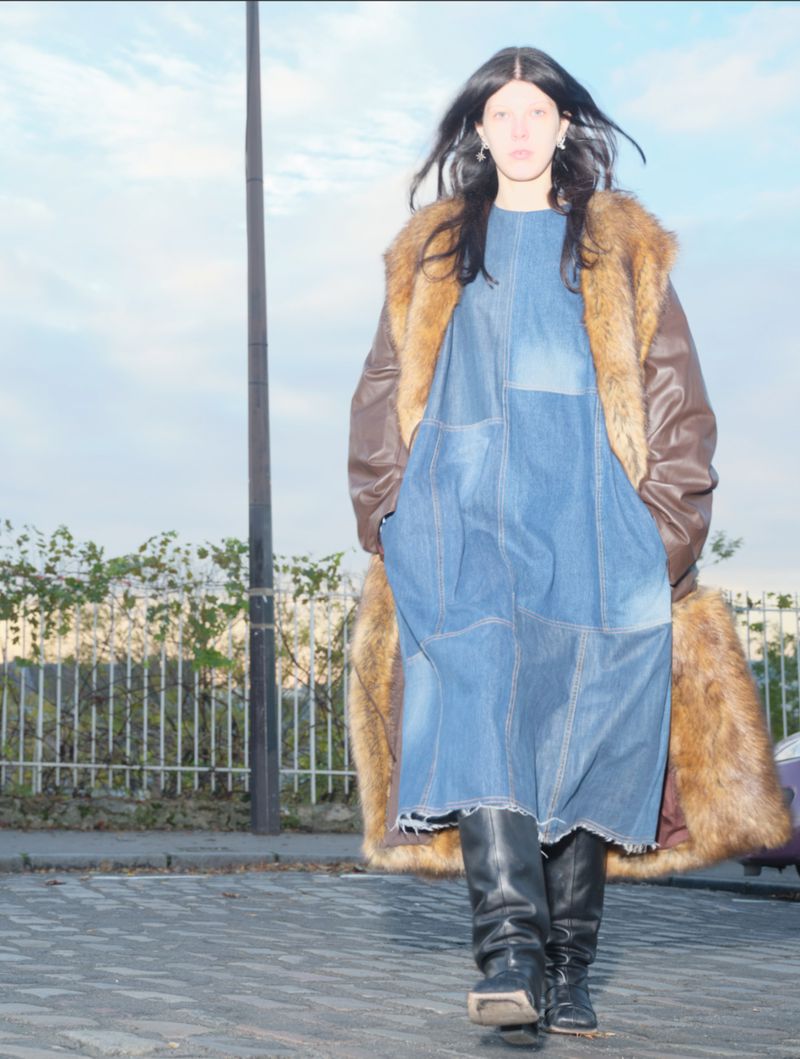 An adult woman stands outside wearing a blue denim dress, brown fur coat, and black boots against a sky and fence backdrop.