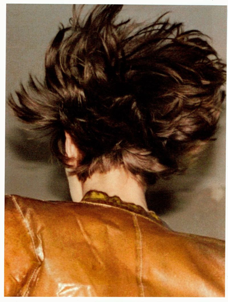 Close-up portrait from behind of a person with dark tousled hair wearing a brown leather jacket against a neutral grey wall.
