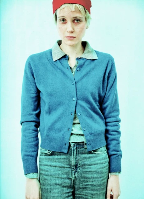 Woman in blue cardigan and jeans with a red head covering, posing in front of a pale blue backdrop in a brightly lit photo studio.