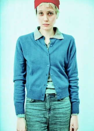 Woman in blue cardigan and jeans with a red head covering, posing in front of a pale blue backdrop in a brightly lit photo studio.