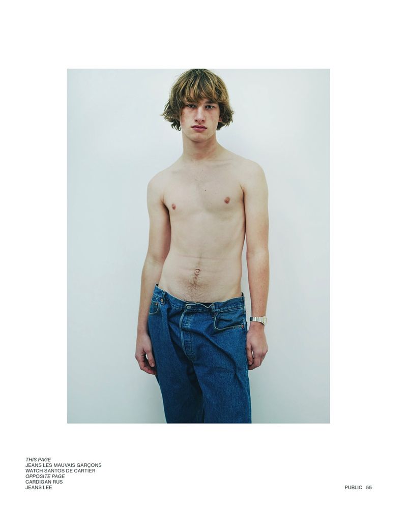 Shirtless male model with blond hair poses in blue jeans against a white wall for a fashion editorial photo in a minimalist style.