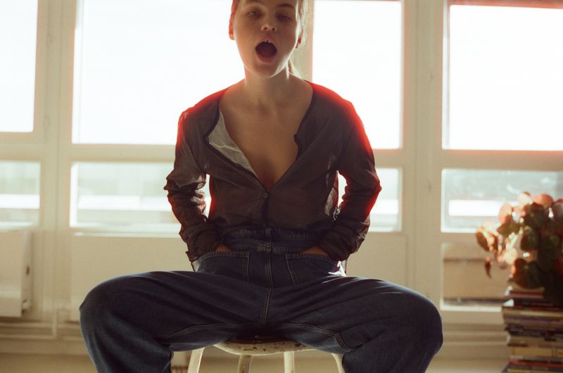 Young adult female model poses in an open-collared shirt and jeans, seated on a chair in a bright room with a neutral background.