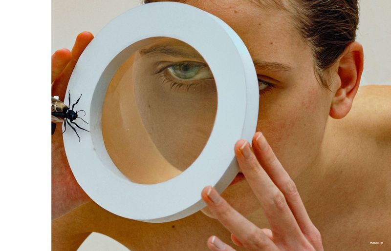 Close-up of a fair-skinned person with green eyes looking through a white circular frame with an insect attached to the side.