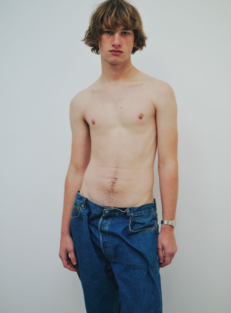 Male fashion model stands shirtless in blue jeans, showcasing a casual yet stylish look against a plain white backdrop.