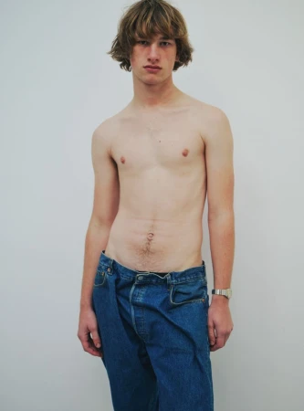 Male fashion model stands shirtless in blue jeans, showcasing a casual yet stylish look against a plain white backdrop.
