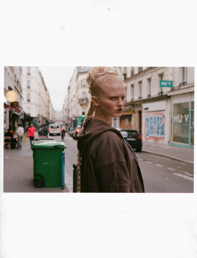 Fashion portrait of a woman with braided blonde hair wearing a brown hooded jacket, standing on a city street in front of buildings.