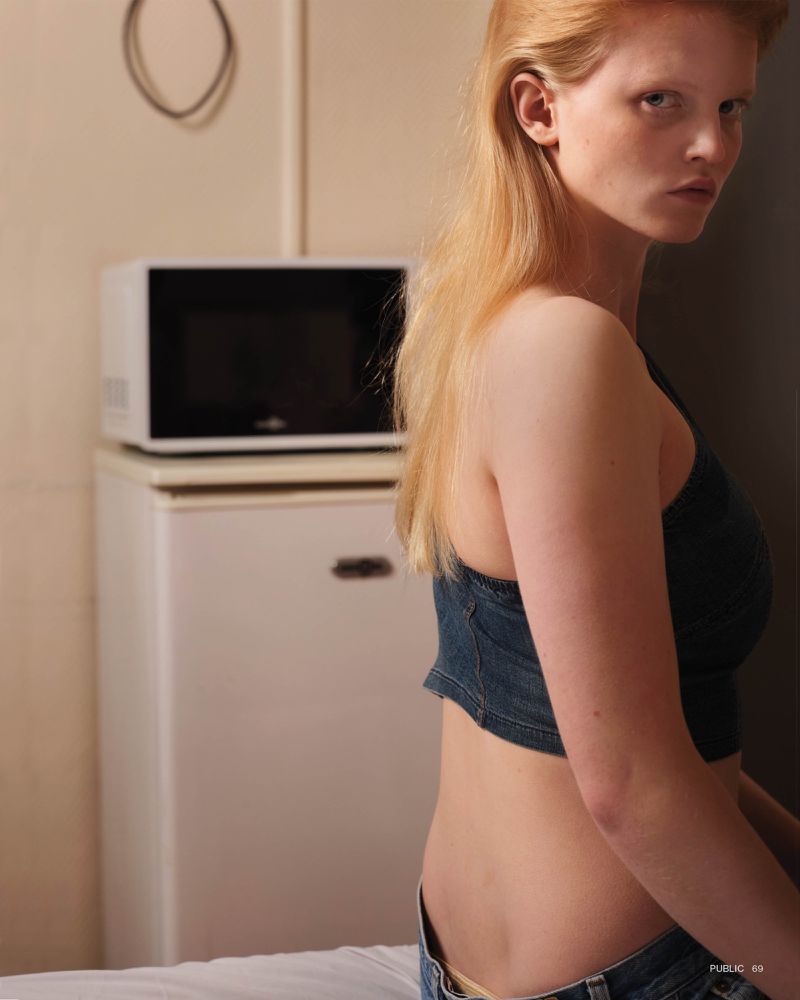 Blonde woman poses indoors wearing a denim top, jeans, and neutral makeup against a backdrop with a mini fridge and microwave.
