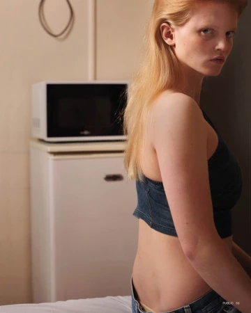 Blonde woman poses indoors wearing a denim top, jeans, and neutral makeup against a backdrop with a mini fridge and microwave.